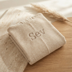 Een gedetailleerde 45-graden productfoto van een beige badcape voor baby's, gepersonaliseerd met de naam 'Sev' in donkerbeige borduring. Liggent op een luxe travertijn en eikenhouten ondergrond met sfeervol licht.