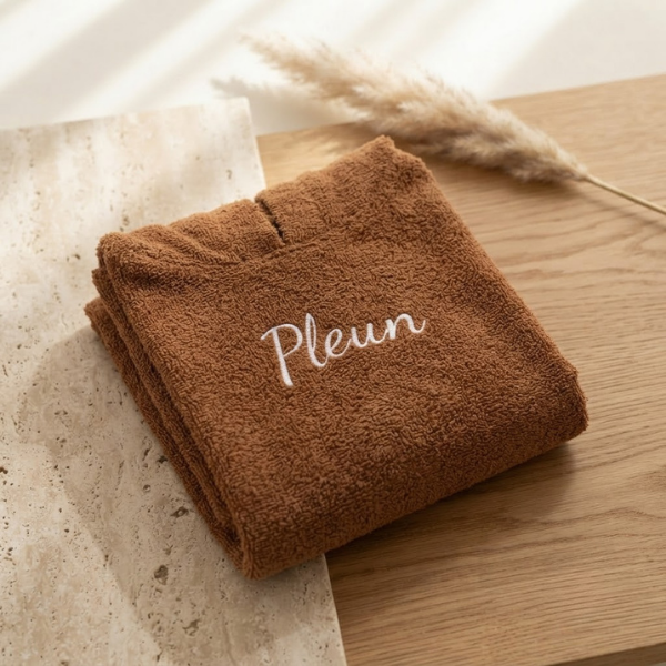Een gedetailleerde 45-graden productfoto van een bruine badcape voor baby's, gepersonaliseerd met de naam 'Pleun' in witte borduring. Liggent op een luxe travertijn en eikenhouten ondergrond met sfeervol licht.