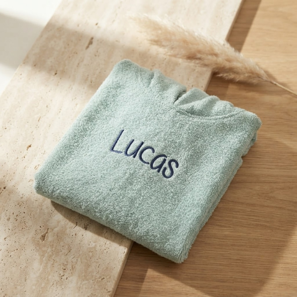 Een gedetailleerde 45-graden productfoto van een mintgroene badcape voor baby's, gepersonaliseerd met de naam 'Lucas' in donkerblauwe borduring. Liggent op een luxe travertijn en eikenhouten ondergrond met sfeervol licht.
