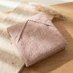Een gedetailleerde 45-graden productfoto van een roze badcape voor baby's, gepersonaliseerd met de naam 'Fien' in beige borduring. Liggent op een luxe travertijn en eikenhouten ondergrond met sfeervol licht.