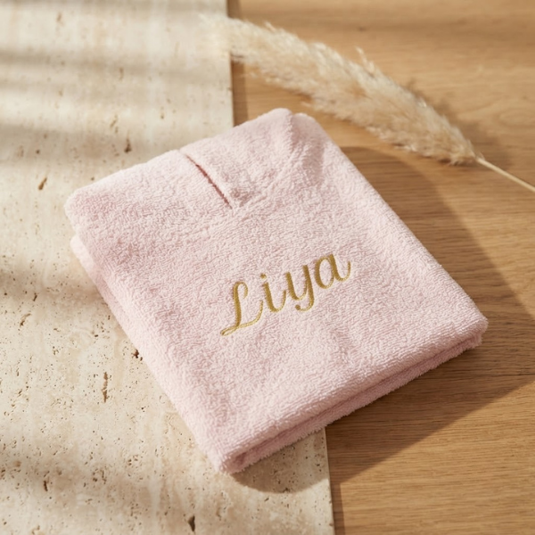 Een gedetailleerde 45-graden productfoto van een roze badcape voor baby's, gepersonaliseerd met de naam 'Liya' in goudkleurige borduring. Liggent op een luxe travertijn en eikenhouten ondergrond met sfeervol licht.