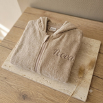 Een gedetailleerde 45-graden productfoto van een beige badjasje voor baby's, gepersonaliseerd met de naam 'Nova' in donkerbruine borduring. Liggent op een luxe travertijn en eikenhouten ondergrond met sfeervol licht.