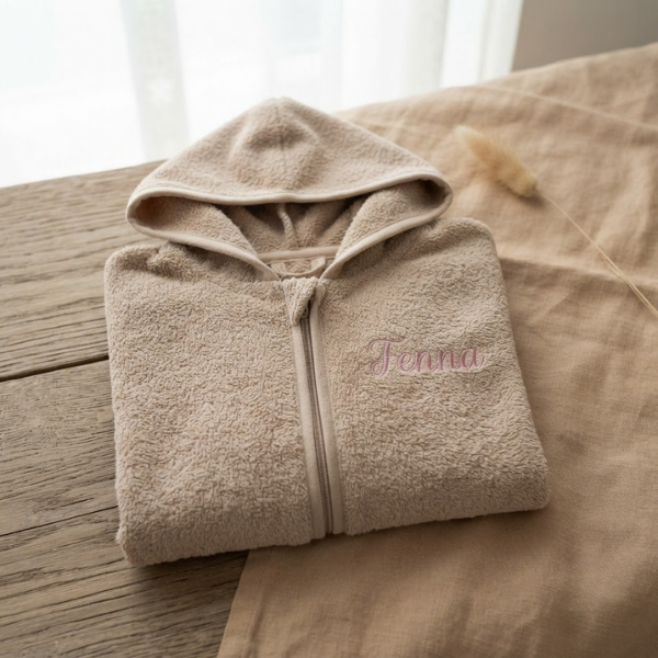 Een gedetailleerde 45-graden productfoto van een beige badjasje met rits, gepersonaliseerd met de naam 'Fenna' in roze borduring. Liggend op een eikenhouten en linnen ondergrond met sfeervol licht.