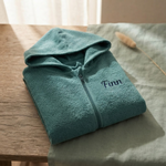 Een gedetailleerde 45-graden productfoto van een lake green babybadjasje met rits, gepersonaliseerd met de naam 'Finn' in donkerblauwe borduring. Liggend op een eikenhouten en linnen ondergrond met sfeervol licht.