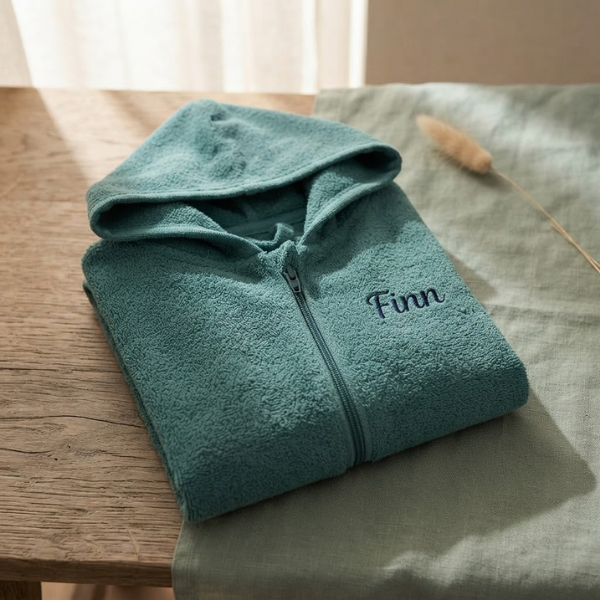 Een gedetailleerde 45-graden productfoto van een lake green babybadjasje met rits, gepersonaliseerd met de naam 'Finn' in donkerblauwe borduring. Liggend op een eikenhouten en linnen ondergrond met sfeervol licht.
