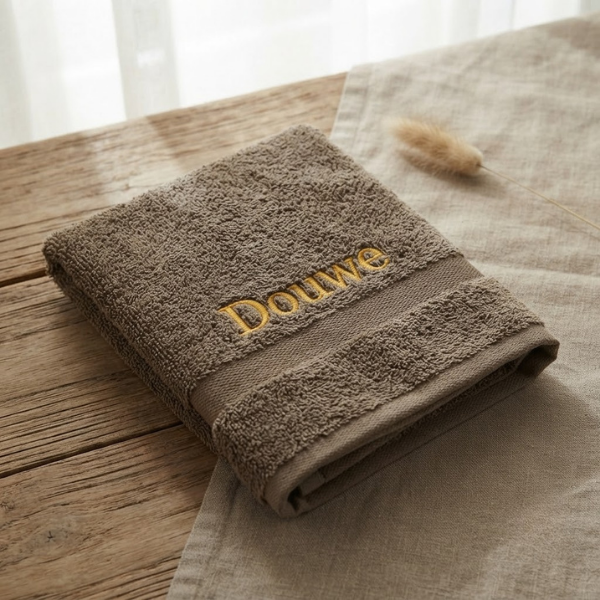 Een gedetailleerde 45-graden productfoto van een bruine Funnies handdoek, gepersonaliseerd met de naam 'Douwe' in goudgele borduring. Liggend op een eikenhouten en linnen ondergrond met sfeervol licht.