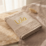 Een gedetailleerde 45-graden productfoto van een beige handdoek, gepersonaliseerd met de naam 'Lola' in gele borduring. Liggend op travertijnsteen en eikenhout met sfeervol licht.