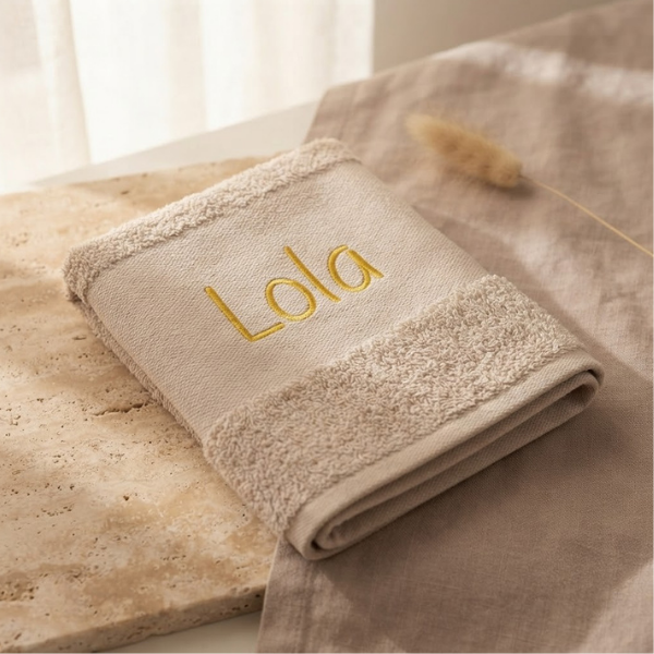 Een gedetailleerde 45-graden productfoto van een beige handdoek, gepersonaliseerd met de naam 'Lola' in gele borduring. Liggend op travertijnsteen en eikenhout met sfeervol licht.