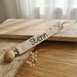 Een gedetailleerde 45-graden productfoto van een beige speenkoord met 'Stenn' borduring, liggend op een combinatie van travertijn steen en eikenhout.