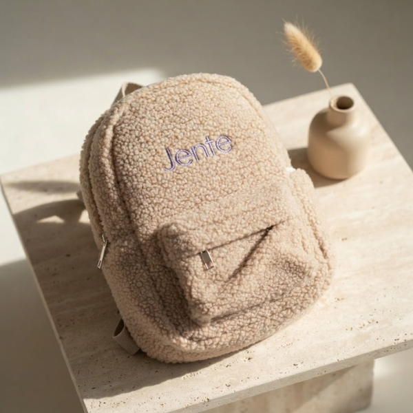 Een individueel productshot van de beige teddy fleece mini backpack 'Jente' met paarse borduring, op een travertijn ondergrond.