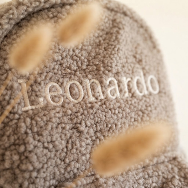 Extreme macro close-up van de beige borduring van de naam 'Leonardo' op de taupe teddy stof, met zichtbare garenstructuur en stofvezels.