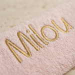 Een macrofoto zoomt in op de goudkleurige borduring van de naam 'Milou' op de roze badstof van het babycapeje. De textuur van het garen en de stof is extreem scherp zichtbaar.