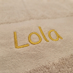 Een macrofoto zoomt in op de gele borduring van de naam 'Lola' op de beige badstof van de handdoek. De textuur van het garen en de lusjes van de stof zijn extreem scherp zichtbaar.