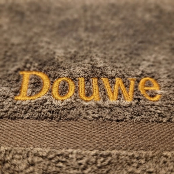 Een macrofoto zoomt in op de goudgele borduring van de naam 'Douwe' op de bruine badstof van de handdoek. De textuur van het garen en de lusjes van de badstof zijn extreem scherp zichtbaar.