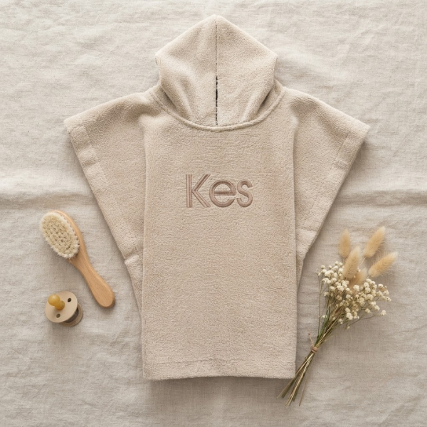 Een stijlvolle flat-lay foto van bovenaf. Het beige badcapeje met 'Kes' borduring ligt op linnen, omringd door een houten babyborstel, een beige speen en een bosje droogbloemen.