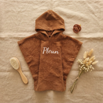 Een stijlvolle flat-lay foto van bovenaf. Het bruine badcapeje met 'Pleun' borduring in wit ligt op linnen, omringd door een houten babyborstel, een terra speen en een bosje droogbloemen.