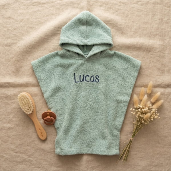 Een stijlvolle flat-lay foto van bovenaf. Het mintgroene badcapeje met 'Lucas' borduring in donkerblauw ligt op linnen, omringd door een houten babyborstel, een terra speen en een bosje droogbloemen.