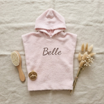 Een stijlvolle flat-lay foto van bovenaf. Het roze badcapeje met 'Belle' borduring in donkerbruin ligt op linnen, omringd door een houten babyborstel, een beige speen en een bosje droogbloemen.