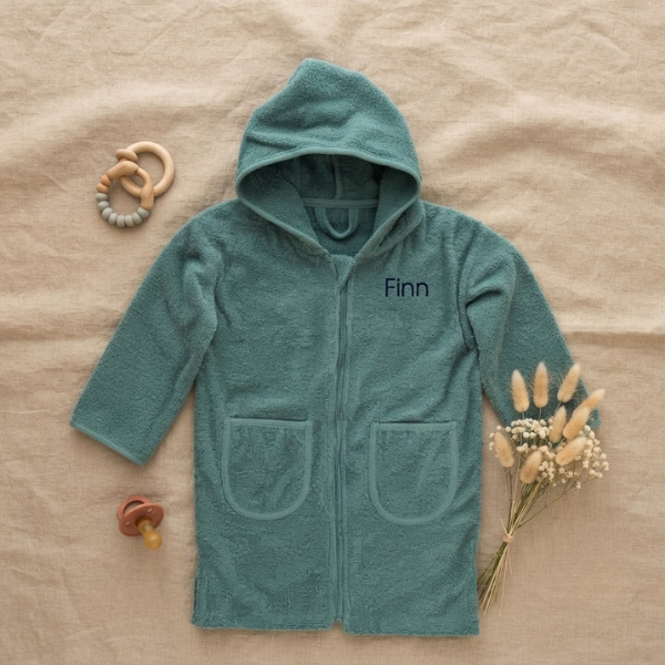 Een stijlvolle flat-lay foto van bovenaf. Het lake green badjasje met 'Finn' borduring in donkerblauw ligt op linnen, omringd door een houten rammelaar, een terra speen en een bosje droogbloemen.