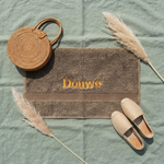 Een stijlvolle flat-lay foto van bovenaf. Het bruine Funnies strandlaken met 'Douwe' borduring in goudgeel ligt op saliegroen linnen, omringd door een rotan tas, espadrilles en pampasgras.