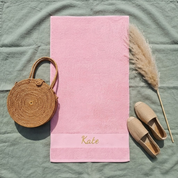 Een stijlvolle flat-lay foto van bovenaf. Het roze Funnies strandlaken met 'Kate' borduring in goudgeel ligt op saliegroen linnen, omringd door een rotan tas, espadrilles en pampasgras.