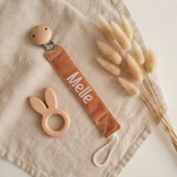 Stijlvolle flat-lay compositie van het cognac velvet speenkoord met witte naam 'Melle', omringd door een houten bijtring en droogbloemen op linnen