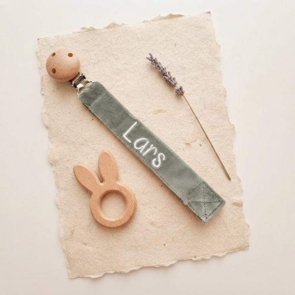 Stijlvolle flat-lay compositie van het saliegroene velvet speenkoord met witte naam 'Lars', omringd door een houten bijtring en gedroogde lavendel op handgeschept papier.
