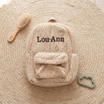 Een bovenaanzicht van de gepersonaliseerde beige teddy rugzak 'Lou-Ann' (donkerbruin garen), omringd door een houten borstel en een saliegroene bijtring op een jute ondergrond.