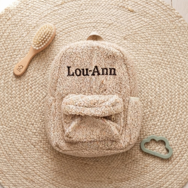 Een bovenaanzicht van de gepersonaliseerde beige teddy rugzak 'Lou-Ann' (donkerbruin garen), omringd door een houten borstel en een saliegroene bijtring op een jute ondergrond.