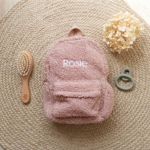 Een bovenaanzicht van de gepersonaliseerde dusty pink teddy rugzak 'Rosie', omringd door een houten borstel, saliegroene bijtring en een droogbloem op een jute ondergrond.
