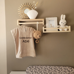 Een sfeerfoto in een babykamer. Het beige badcapeje met de donkerbeige borduring 'Grote broer Abe' hangt aan een houten haakje aan een zandkleurige muur. Op de achtergrond een commode, rotan spiegel en een schapje met een Nijntje knuffel.