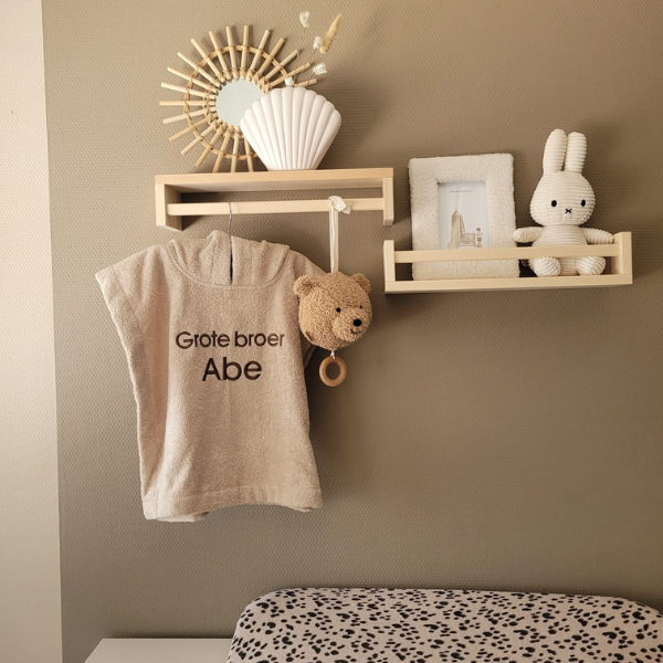 Een sfeerfoto in een babykamer. Het beige badcapeje met de donkerbeige borduring 'Grote broer Abe' hangt aan een houten haakje aan een zandkleurige muur. Op de achtergrond een commode, rotan spiegel en een schapje met een Nijntje knuffel.