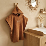 Een sfeerfoto in een babykamer. Het bruine badcapeje met de witte borduring 'Senn' hangt aan een houten haakje aan een zandkleurige muur. Op de achtergrond een commode, rotan spiegel en een schapje met een teddybeer.