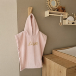 Een sfeerfoto in een babykamer. Het roze badcapeje met de goudkleurige borduring 'Liya' hangt aan een houten haakje aan een zandkleurige muur. Op de achtergrond een commode, rotan spiegel en een schapje met droogbloemen.