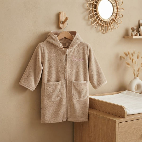 Een sfeerfoto in een babykamer. Het beige badjasje met de roze borduring 'Fenna' hangt aan een houten haakje aan een zandkleurige muur. Op de achtergrond een commode, rotan spiegel en een schapje met een teddybeer.