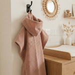 Een sfeerfoto in een moderne badkamer. Het roze badcapeje met de beige borduring 'Fien' hangt aan een matzwart haakje aan een tegelwand. Op de achtergrond een houten badmeubel en hamamdoek.