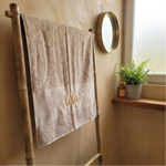 Een sfeerfoto in een rustige badkamer. De beige handdoek met de gele borduring 'Lola' hangt aan een houten bamboe rekje. Op de achtergrond een tadelakt muur en messing spiegel.