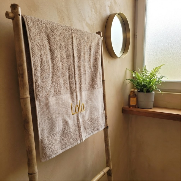 Een sfeerfoto in een rustige badkamer. De beige handdoek met de gele borduring 'Lola' hangt aan een houten bamboe rekje. Op de achtergrond een tadelakt muur en messing spiegel.