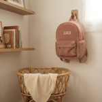 De gepersonaliseerde roze teddy rugzak 'Julia' hangend aan een eikenhouten haakje in een sfeervolle babykamer, met een rotan wiegje op de achtergrond.