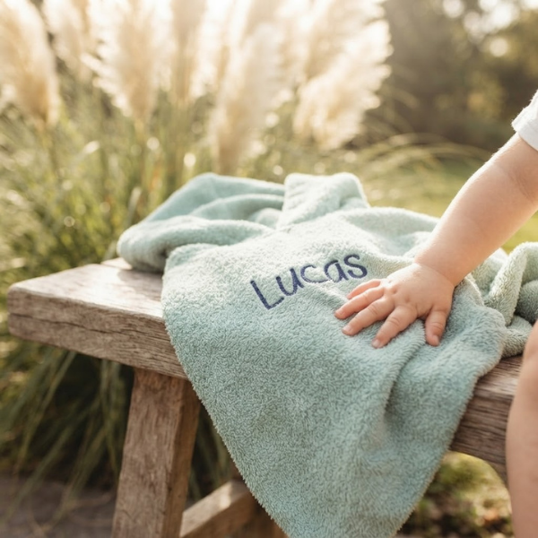 Een lifestylefoto buiten in een tuin. Op een houten bankje ligt het mintgroene badcapeje met donkerblauwe borduring 'Lucas'. Een zacht babyhandje ligt op de badstof. Op de achtergrond is wapperend pampasgras te zien.