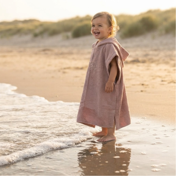 Een lifestylefoto buiten op het strand. Een peuter draagt het roze badcapeje met beige borduring 'Fien' en staat met de voetjes in het zeewater aan de vloedlijn.