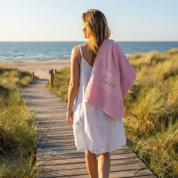 Een lifestylefoto buiten in de duinen. Een vrouw met een witte jurk loopt over een houten pad. Ze draagt het roze Funnies strandlaken met de goudgele borduring 'Kate' over haar schouder. Helmgras op de achtergrond.