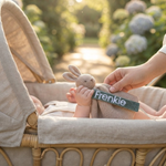 Een babyhandje houdt een knuffel vast waaraan het saliegroene velvet speenkoord met witte borduring 'Frenkie' is bevestigd, in een zonovergoten kinderwagen.
