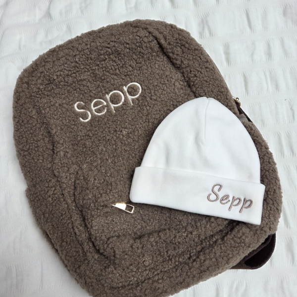 Een kraamcadeau voor Sepp bestaande uit een taupe kleurige kinderrugzak met naam gepersonaliseerd en witte kindermuts. 