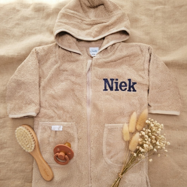 Een stijlvolle flat-lay foto van bovenaf. Het beige badjasje met 'Niek' borduring in donkerblauw ligt op linnen, omringd door een houten babyborstel, een terra speen en een bosje droogbloemen.