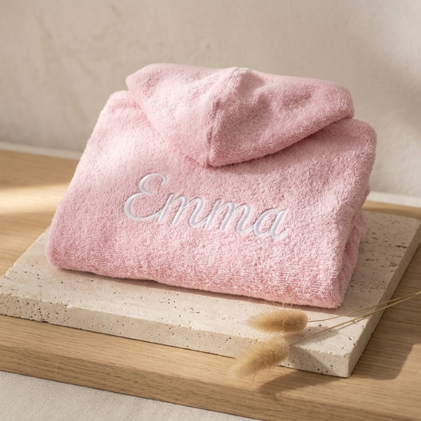 Een schuin bovenaanzicht van de rugzijde van het roze babybadjasje met de naam 'Emma' in wit sierlijk borduurwerk, gepresenteerd op een travertijn blok op eikenhout.