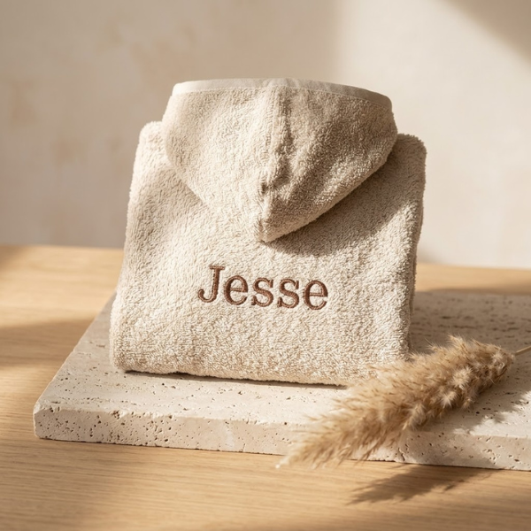 Een schuin bovenaanzicht van de rugzijde van een zandkleurige babybadjas, waarop de naam 'Jesse' in bruin garen is geborduurd, gepresenteerd op travertijn en eikenhout.