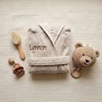 Een bovenaanzicht (flat-lay) van de zandkleurige babybadjas met de naam 'Lennon', omringd door een houten borstel, een speen en een teddy knuffel op een crème ondergrond.