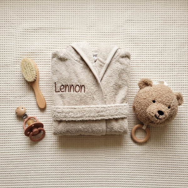 Een bovenaanzicht (flat-lay) van de zandkleurige babybadjas met de naam 'Lennon', omringd door een houten borstel, een speen en een teddy knuffel op een crème ondergrond.