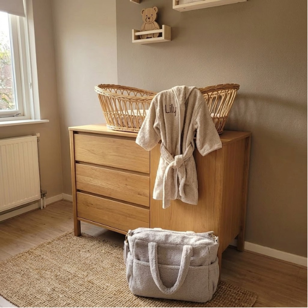 Een sfeervolle opname in een babykamer interieur, waar de gepersonaliseerde 'Lily' badjas over een rotan mozesmand op een eikenhouten commode hangt.